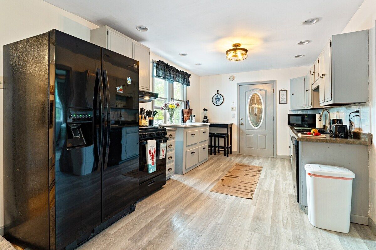 A modern kitchen with a black refrigerator, white cabinets, a wooden floor, and a small trash bin.
