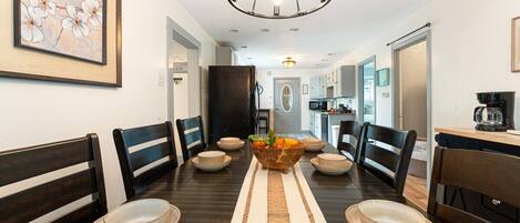 A dining area with a long table, chairs, a central fruit bowl, and a chandelier.