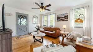 Una habitación con ventilador de techo, un sofá marrón, un sillón blanco, una mesita y una lámpara.