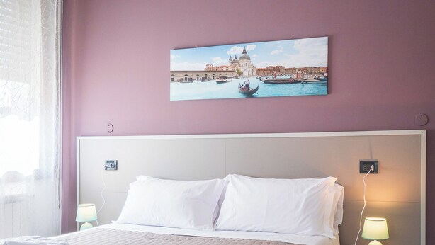 Habitación de hotel con cama, almohadas blancas, mesita de noche con lámpara y un cuadro de un paisaje urbano en la pared.