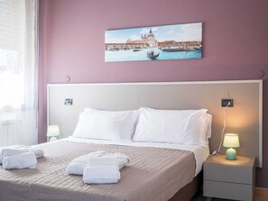 Una camera d'albergo con un letto, cuscini bianchi, un comodino con una lampada e un quadro che raffigura un panorama urbano.