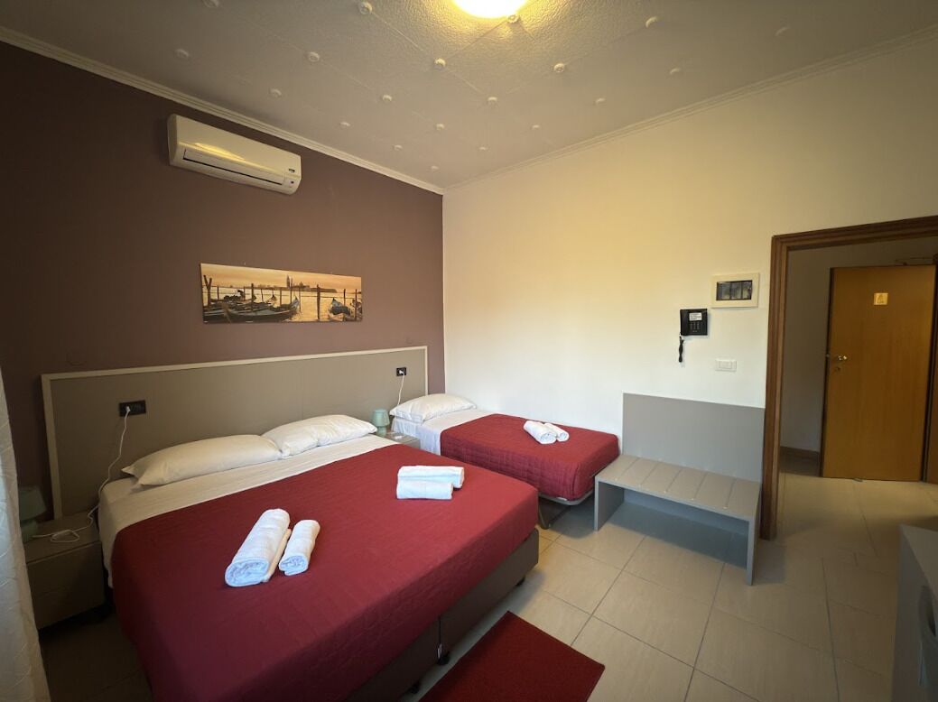 A hotel room with two beds, a bench, an air conditioning unit, and a painting on the wall.