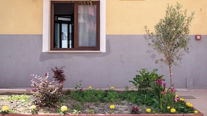 Entrada de un hotel con ventana, un pequeño balcón y un jardín con diversas plantas y flores.