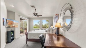Een slaapkamer met een bed, een bureau, een stoel, een plafondventilator, een grote spiegel en een televisie.