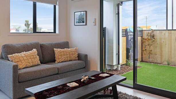 Una sala de estar moderna con un sofá gris, dos cojines, una mesa de centro oscura con libros y vistas a un césped verde a través de puertas de cristal grandes.
