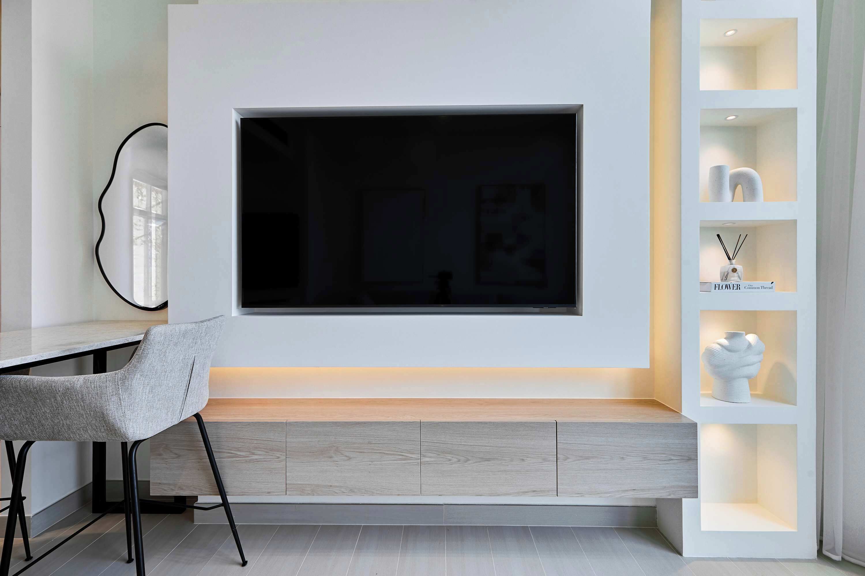 Una sala moderna con un televisor de pantalla plana grande montado en una pared blanca, un sillón tapizado gris, un mueble de madera debajo del televisor y una estantería blanca con objetos decorativos.