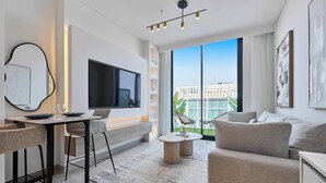 A modern living room with a flat-screen TV, a dining area with bar stools, a sofa, and a large window offering a city view.