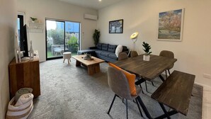 A modern living room with a dark sofa, wooden coffee table, and a dining table with benches. There is a large window providing natural light and a view of the outdoors. - Modern and Contemporary 3BR Home (Christchurch)