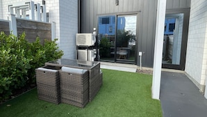 A modern outdoor seating area with wicker furniture and artificial grass, adjacent to a building with a glass door and a grey exterior wall. - Modern and Contemporary 3BR Home (Christchurch)