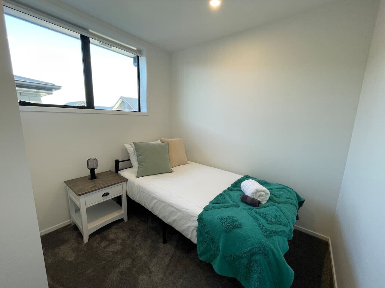 Een kleine, verzorgde slaapkamer met een enkel bed, een nachtkastje en een raam.
