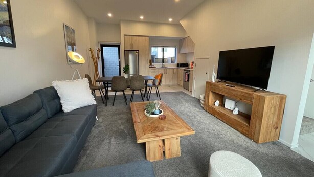 A modern living room with a sofa, coffee table, ottoman, and a TV on a wooden stand. There is a dining area with chairs and a table in the background. - Modern and Contemporary 3BR Home (Christchurch)
