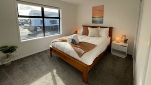 A modern bedroom with a large window, a wooden bed, a bedside table with a lamp, and a view of the outside. - Modern and Contemporary 3BR Home (Christchurch)