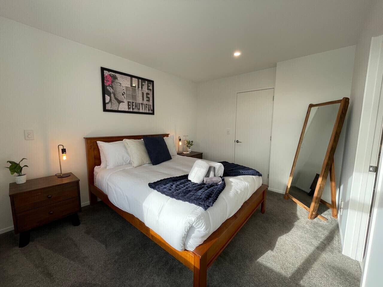 Een keurig opgemaakte slaapkamer met een bed, nachtkastje, lamp en een spiegel.