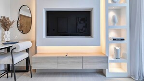 Un salon moderne avec un téléviseur à écran plat fixé à un mur blanc, un banc en bois, une petite table avec un vase et un verre à vin, et une étagère blanche agrémentée d’objets décoratifs.