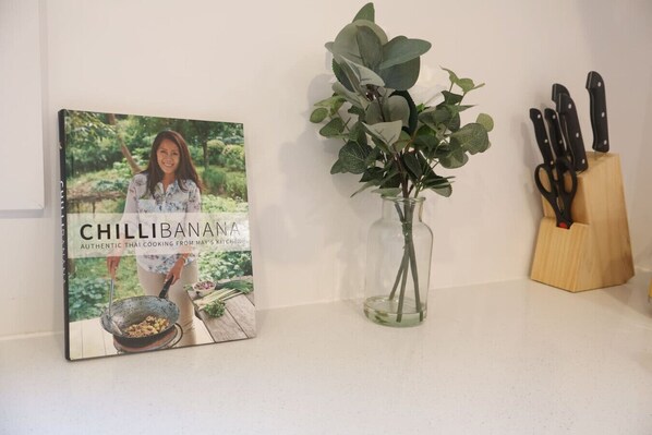 A cookbook titled 'CHILLIBANANA' on a kitchen counter, a vase with green leaves, and a knife block.