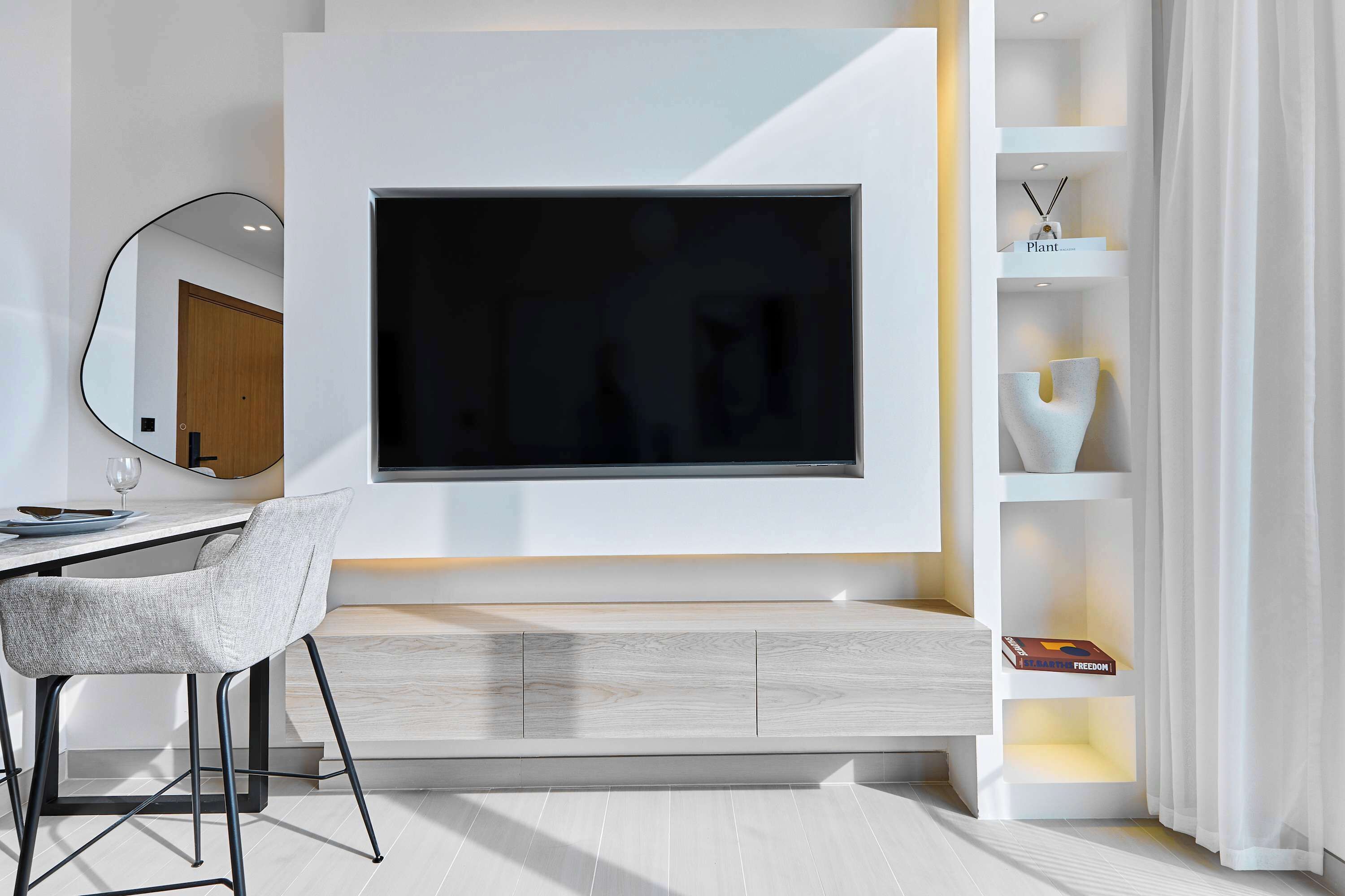 A modern living room with a large flat-screen TV mounted on a white wall, a dining area with a light-colored chair, and a built-in shelf with decorative items.