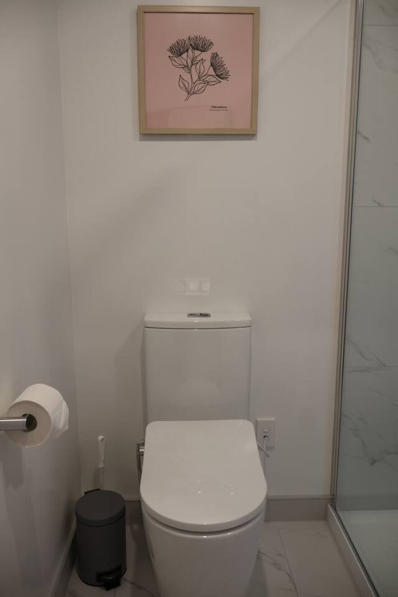 A small bathroom with a toilet, a trash bin, and a framed botanical illustration on the wall.