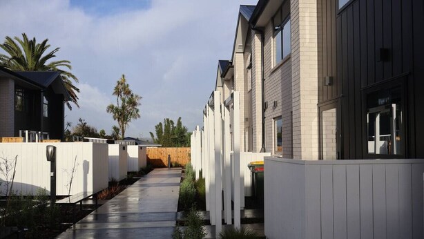 Una zona residenziale moderna con un vialetto in cemento, recinzioni bianche e case con rivestimenti scuri e ampie finestre.