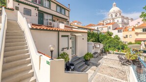 A building with a staircase leading to a balcony, a small outdoor seating area, and a view of a domed building in the background. - GuestReady - Alfama Terrace B1 (Lisbon)