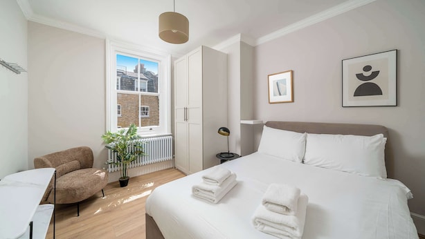 A modern bedroom with a large bed, a chair, a desk, and a window with a view of buildings.