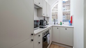 Cocina compacta con armarios blancos, microondas, tostadora y ventana con vista a un edificio de ladrillo.
