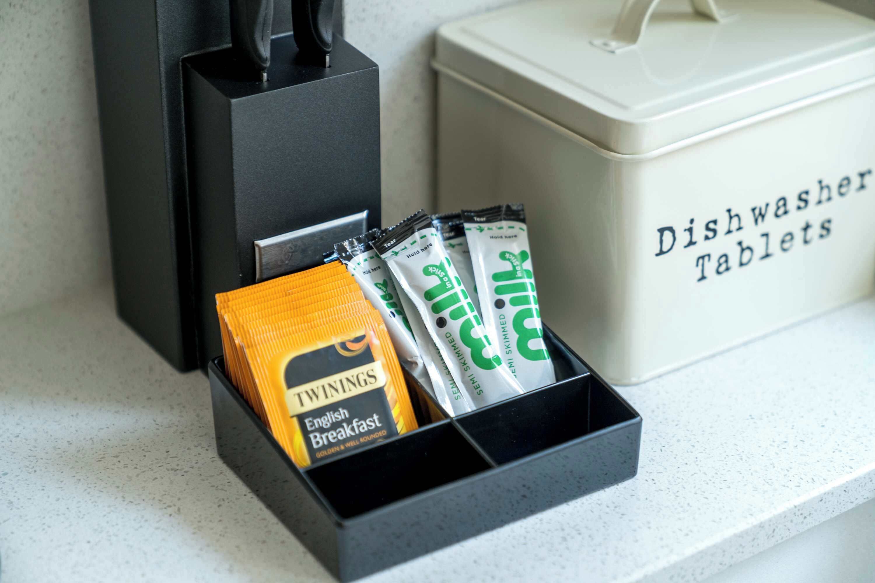 A kitchen counter with a dishwashing tablet dispenser, a coffee maker, and a container labeled "Dishwasher Tablets".