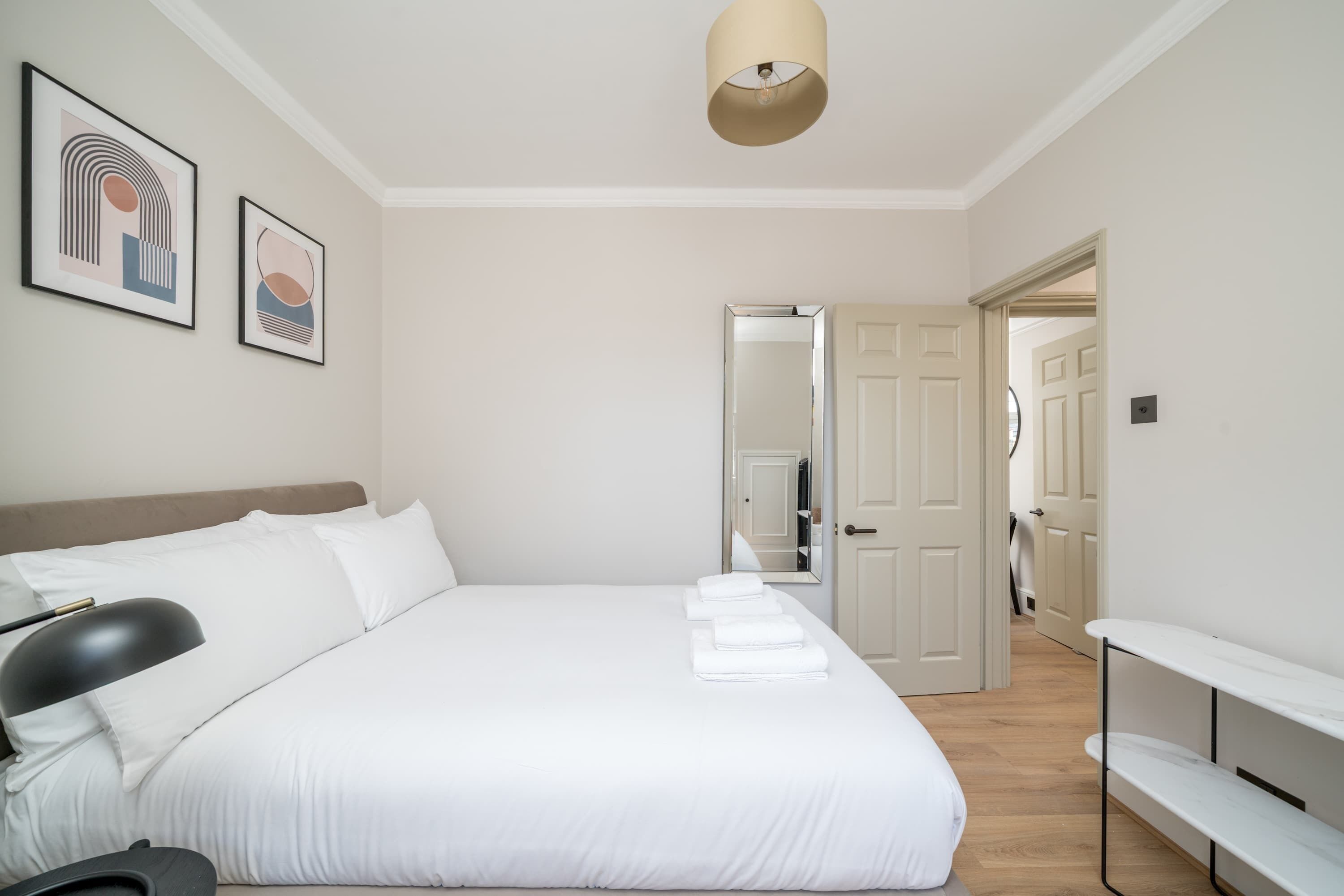 Ein ordentlich eingerichtetes Schlafzimmer mit einem Bett, Nachttischen, einer Lampe und gerahmten Bildern an der Wand.