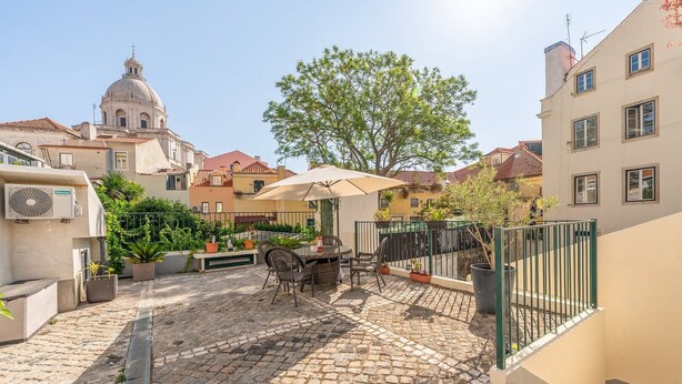 A courtyard with a table and chairs, surrounded by potted plants and a large tree. - GuestReady - Alfama Terrace G (Lisbon)