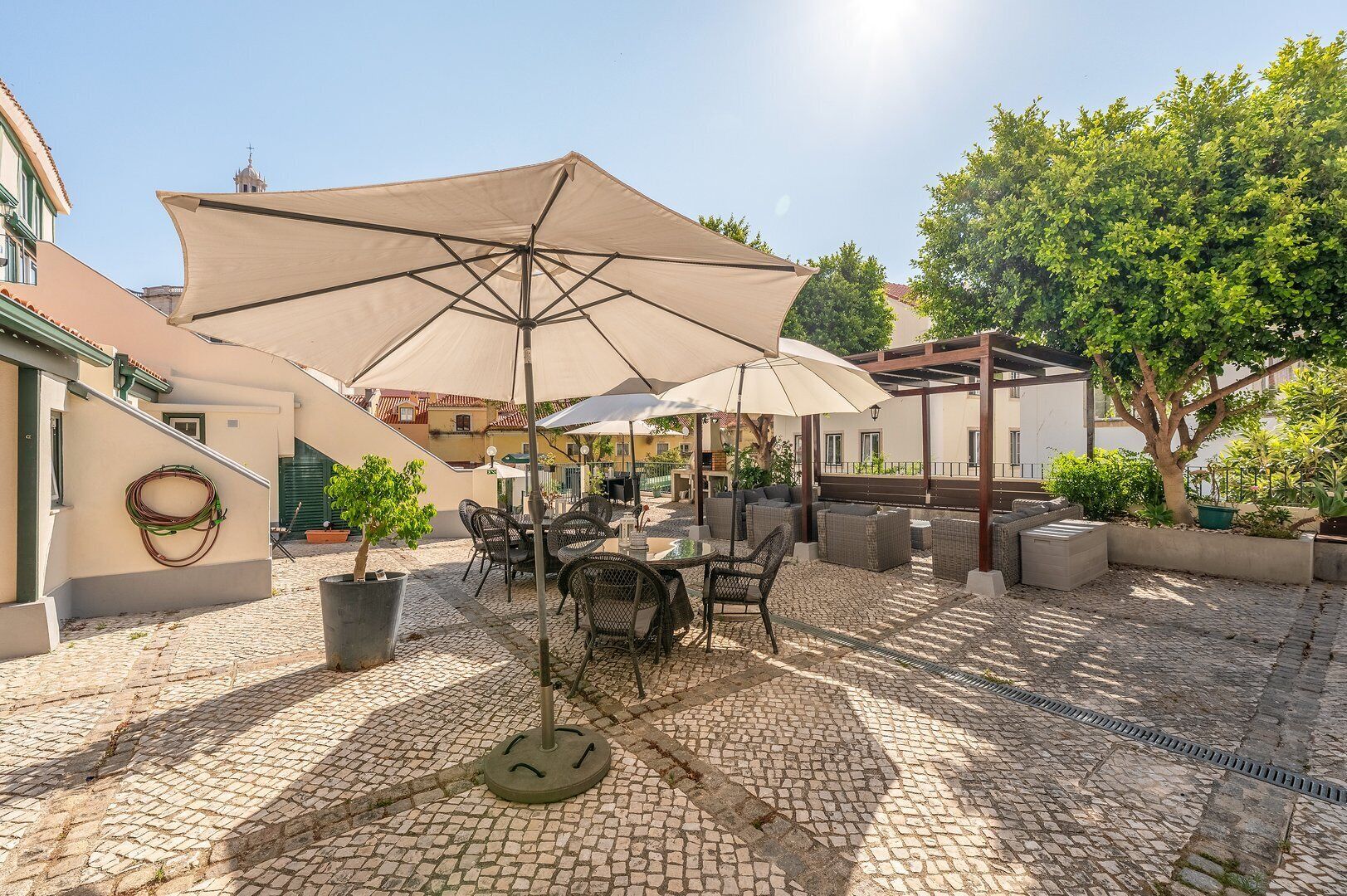 An outdoor patio with tables and chairs under large umbrellas, surrounded by cobblestone flooring and greenery.