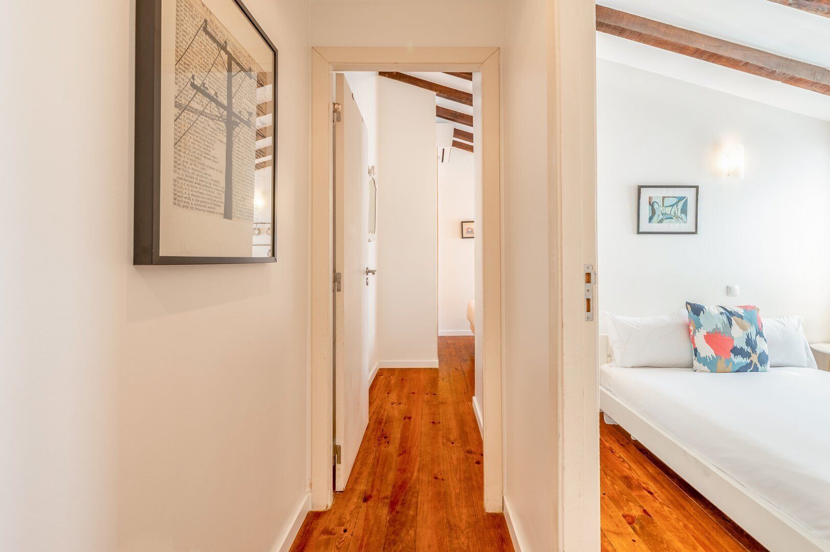 Corredor com piso de madeira que leva a um quarto com cama e luminária na parede.