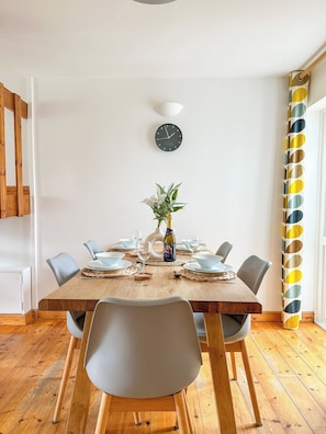 A dining area with a wooden table set for four, surrounded by chairs, a clock on the wall, and a plant centerpiece.