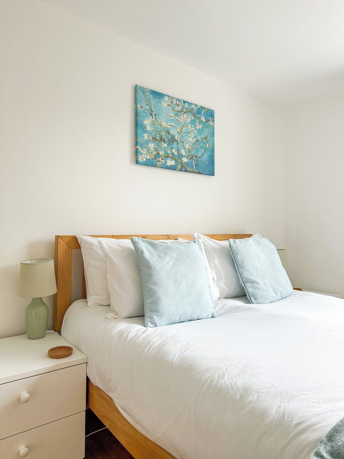 A neatly made bed with white bedding and light blue pillows, a bedside table with a lamp, and a wall-mounted painting of a tree.