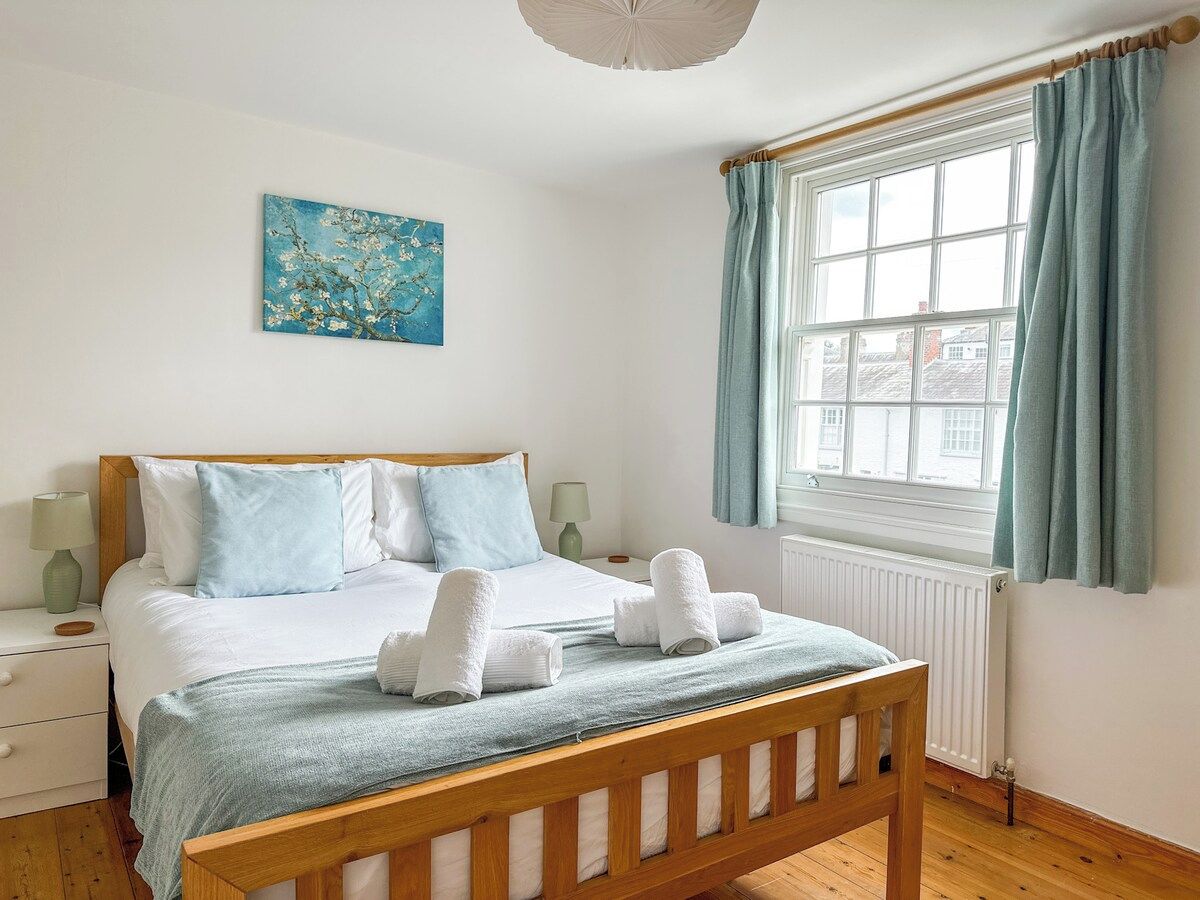 A bedroom with a wooden bed, white bedding, a blue artwork on the wall, and a window with curtains.