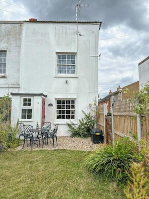 A two-story white building with a red door, a small garden with a table and chairs, and a wooden fence.