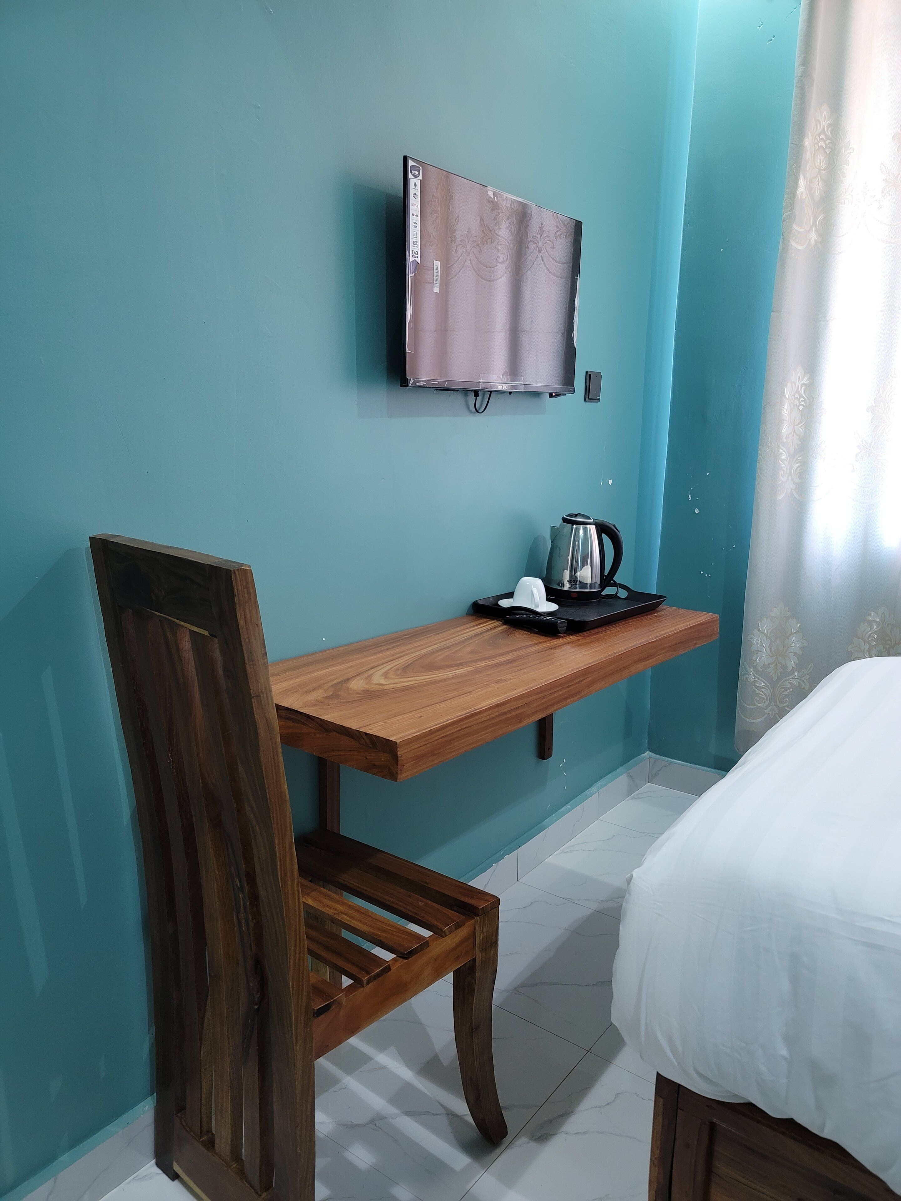 Una habitación con paredes color turquesa, un escritorio de madera con una silla, una cama con ropa de cama blanca, un televisor montado en la pared y una tetera sobre el escritorio.