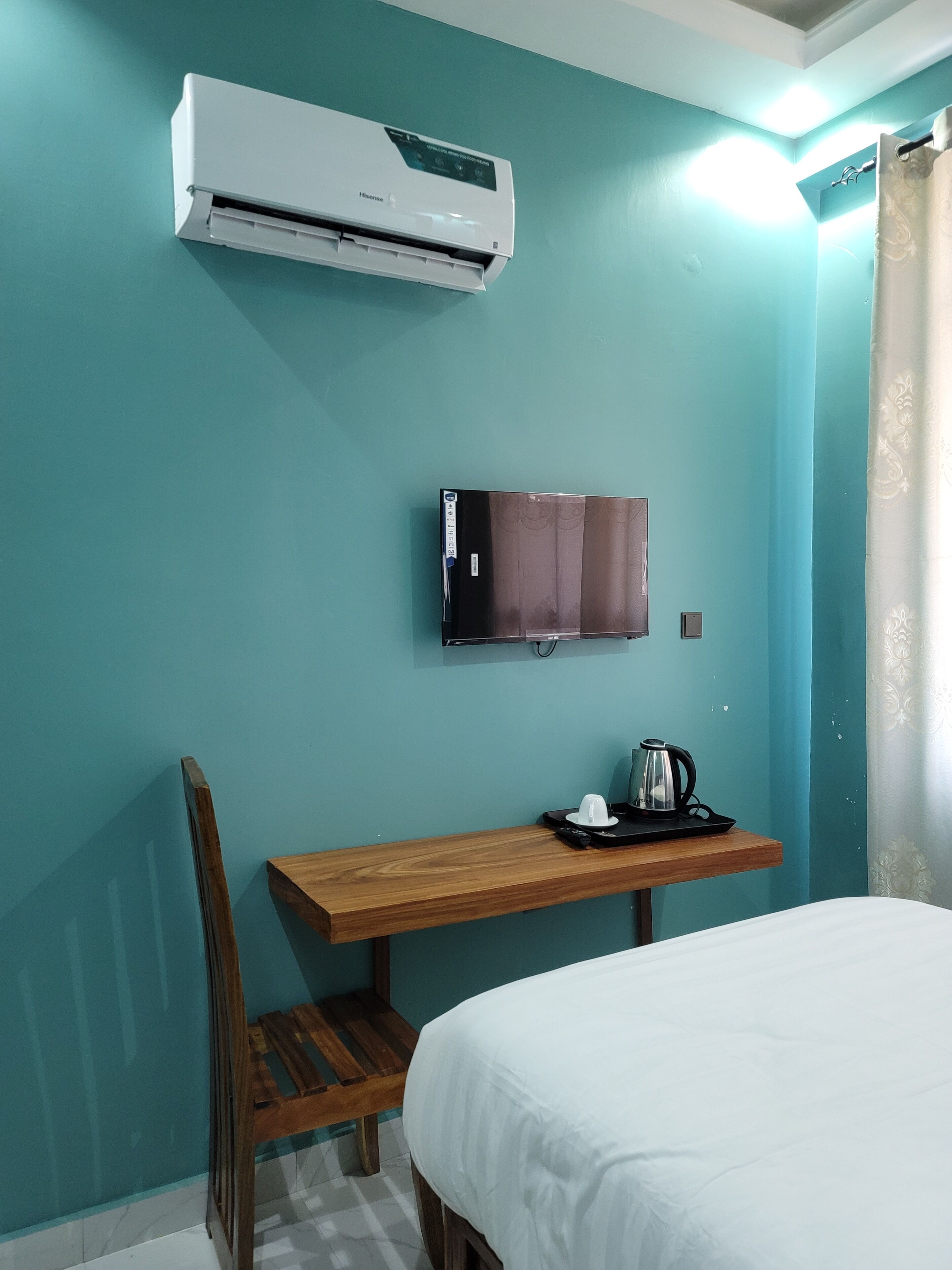 A hotel room with teal walls, a bed, a wooden desk with a chair, a flat-screen TV, and an air conditioner.