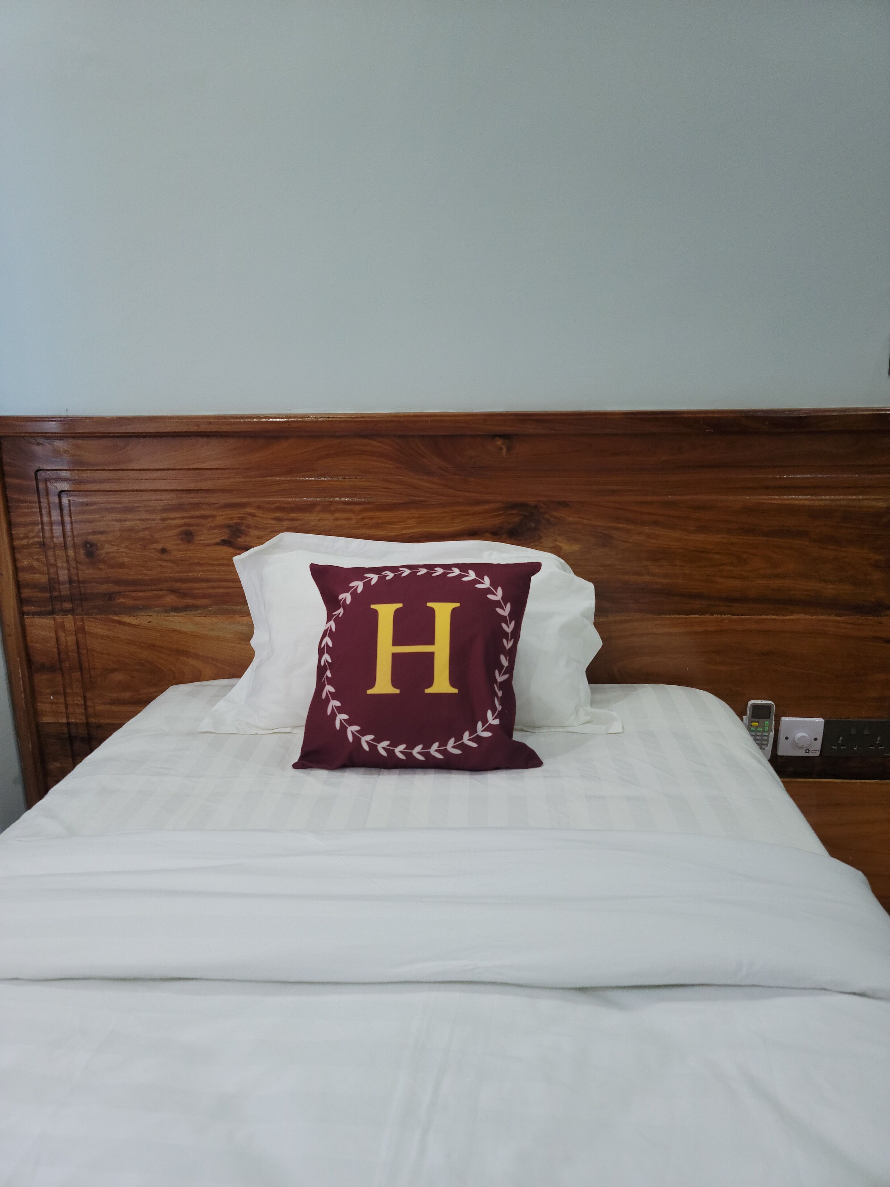 A neatly made bed with white linens, a wooden headboard, and a pillow with a large 'H' and a laurel wreath design.
