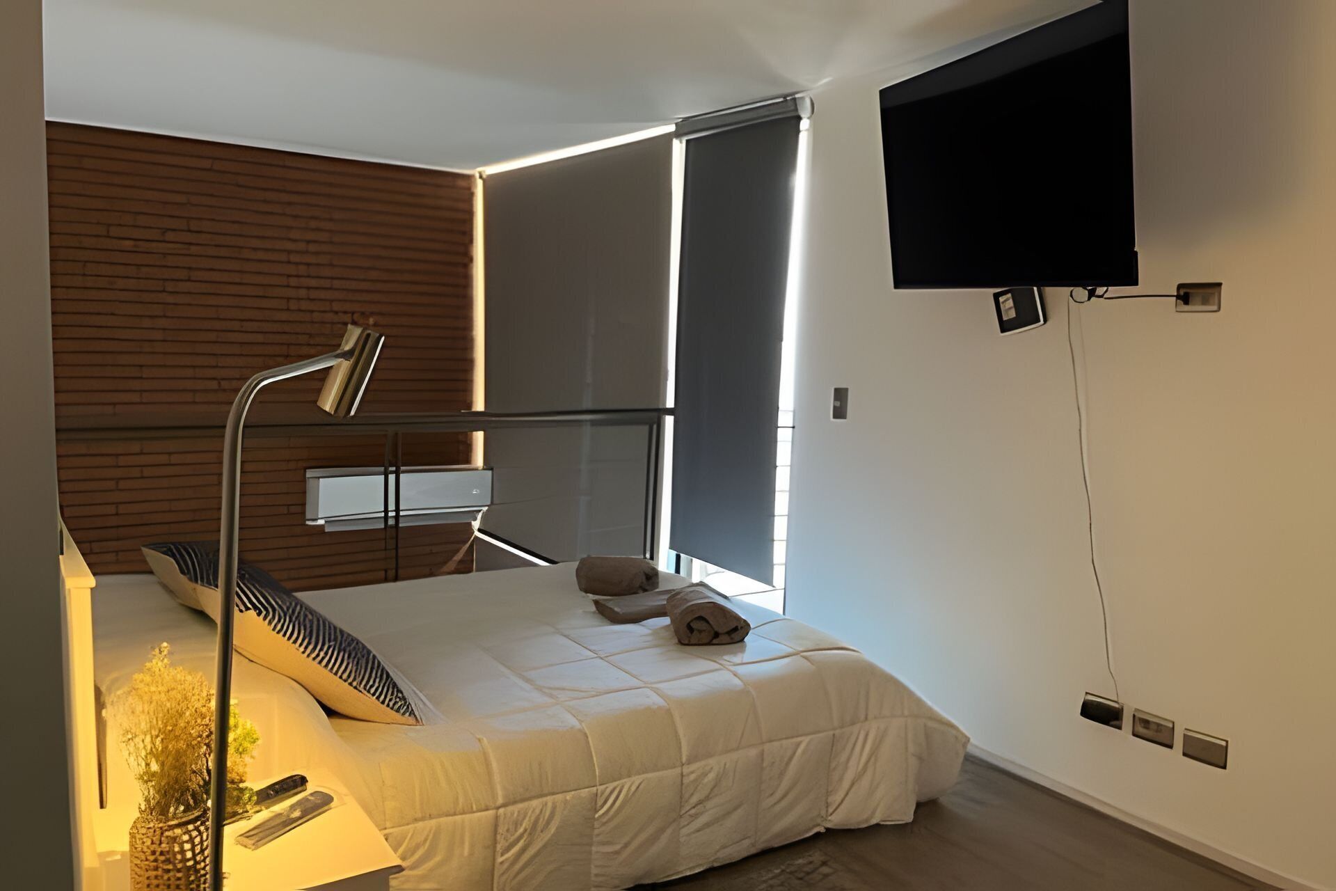 A modern bedroom with a bed, a desk, a chair, a TV mounted on the wall, and a window with blinds.