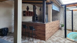 An outdoor kitchen area with a built-in bar, stone walls, and a pool. - House with Heated Pool - Perequê-Açu - UBATUBA - SP (Perequê-Acu Ubatuba)