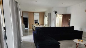 A living room with a black sectional sofa, a small round table with two yellow vases, and a kitchen in the background. - House with Heated Pool - Perequê-Açu - UBATUBA - SP (Perequê-Acu Ubatuba)