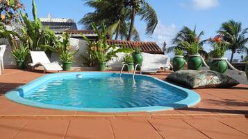 Piscina retangular com borda azul, rodeada por um deck ladrilhado. Há espreguiçadeiras e plantas em vasos ao redor da piscina.