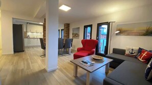 Un salon moderne avec un fauteuil rouge, un canapé gris et une table basse. On aperçoit une cuisine à l’arrière-plan, avec des meubles blancs et un mur en briques apparentes.