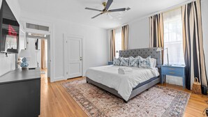 Een slaapkamer met een groot bed, een plafondventilator, een nachtkastje en een raam met gordijnen.