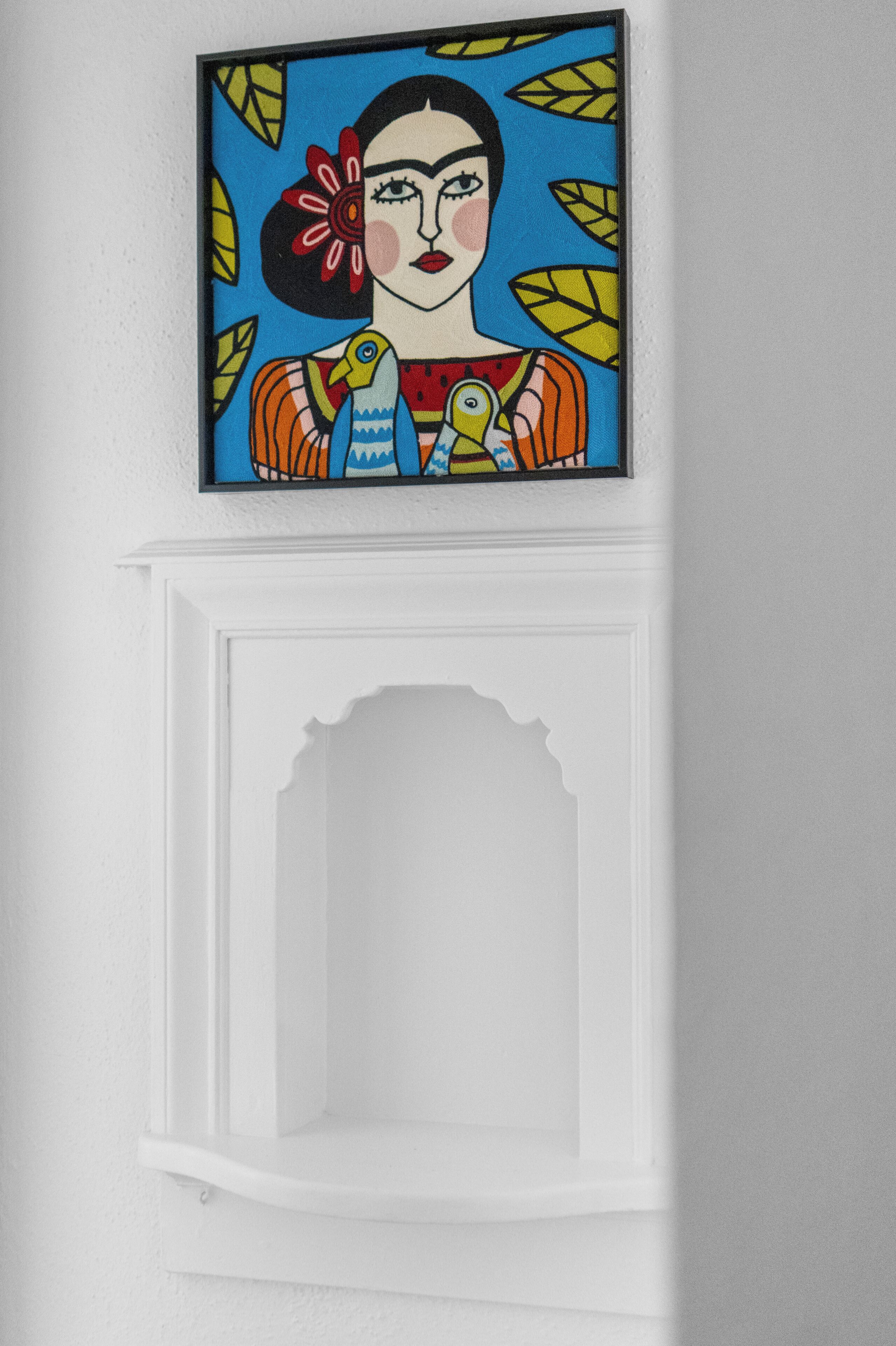 A framed artwork with a woman and bird on a blue background, mounted above a white fireplace mantel.