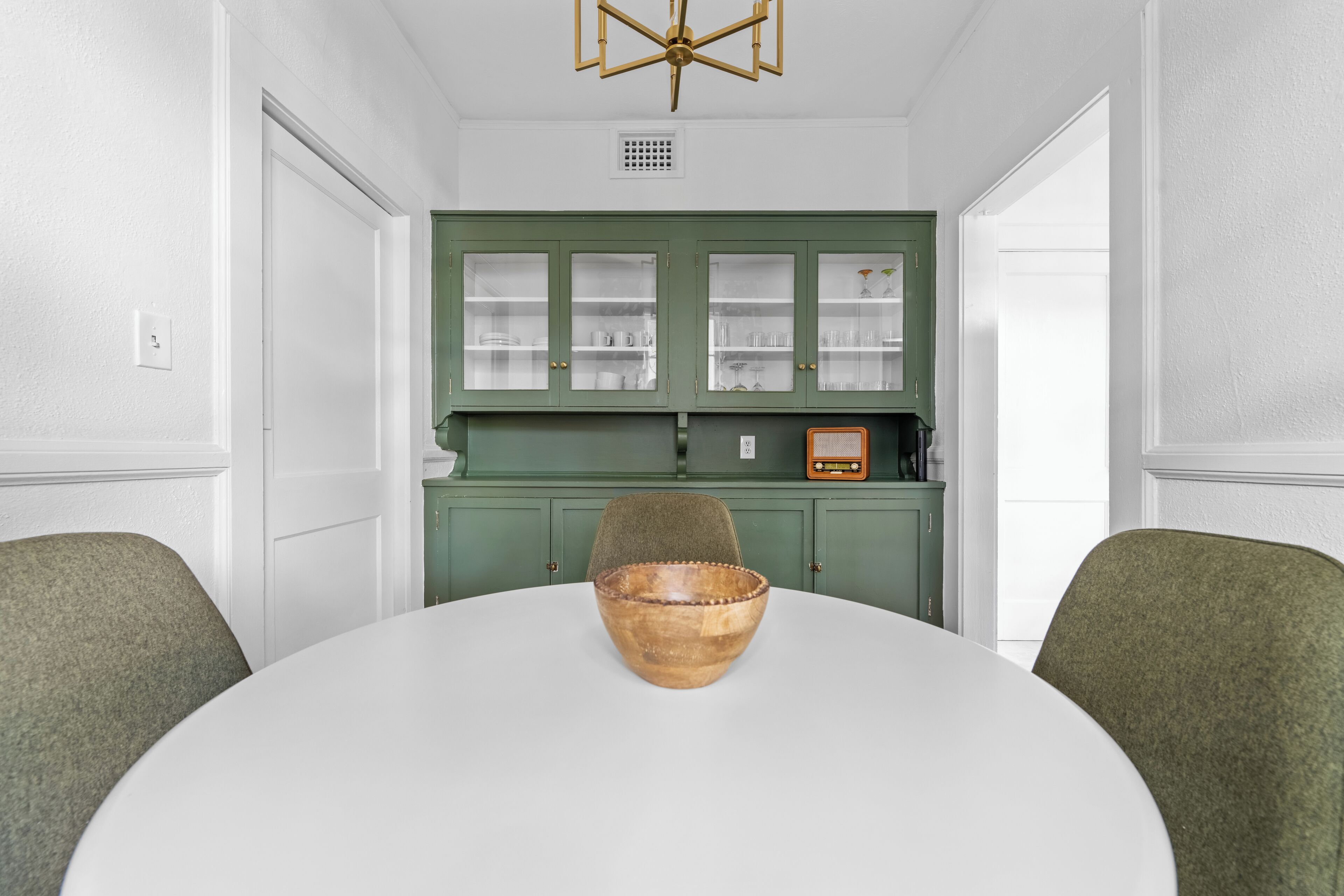 A dining area with a round table, two chairs, a wooden bowl, and green cabinets with glass doors.