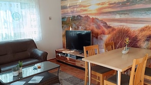 Een woonkamer met een bruine leren bank, een glazen salontafel met planten, een tv op een houten standaard en een eettafel met houten stoelen.