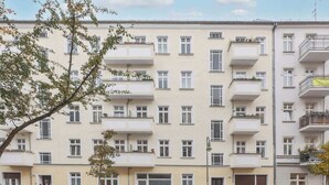 A multi-story residential building with balconies, a parking area with several cars, and a tree in the foreground. - Berlinkiez (Berlin)