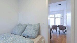 A bedroom with a bed, two pillows, and a bedside table. There is a door leading to a dining area with a table and chairs. - Berlinkiez (Berlin)