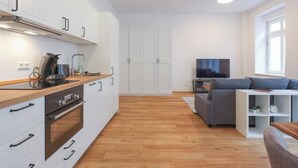 A modern kitchen with white cabinets, a wooden countertop, and a built-in oven. There is a grey sofa and a white TV stand in the living area. - Berlinkiez (Berlin)