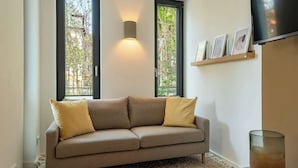 Ein modernes Wohnzimmer mit einem beigefarbenen Sofa, zwei Kissen, einem Glastisch mit Kerze und einem an der Wand befestigten Fernseher.